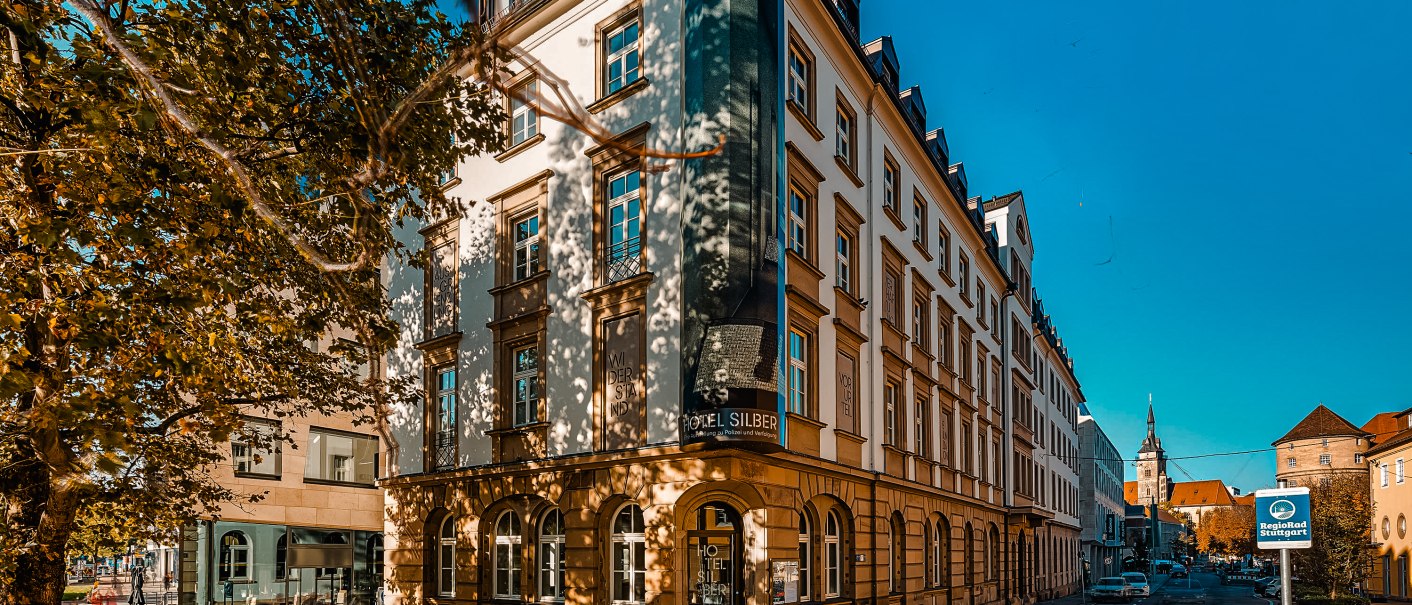 Das Hotel Silber in Stuttgart, ein historisches Gebäude bei sonnigem Wetter, umgeben von Bäumen und einer ruhigen Straße., © Daniel Stauch Photography