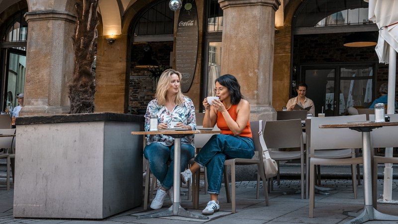 Zwei Frauen sitzen in einem Straßencafé, trinken Kaffee und unterhalten sich. Im Hintergrund sind weitere Gäste und ein Café-Eingang zu sehen., © Stuttgart Marketing GmbH - Sarah Schmid