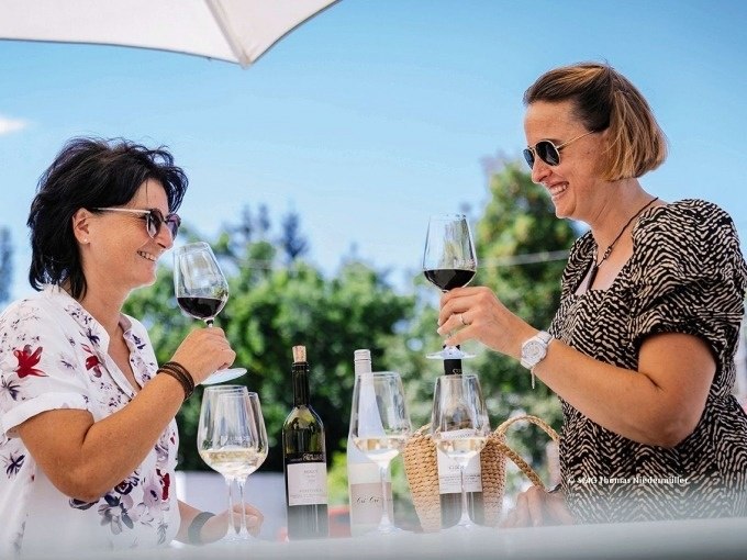 Zwei Frauen stoßen mit Weingläsern an, sitzen unter einem Sonnenschirm, umgeben von Weinflaschen und Gläsern, im Freien., © SMG, Thomas Niedermüller Zwei Frauen stoßen mit Weingläsern an, sitzen unter einem Sonnenschirm, umgeben von Weinflaschen und Gläsern, im Freien., © SMG, Thomas Niedermüller