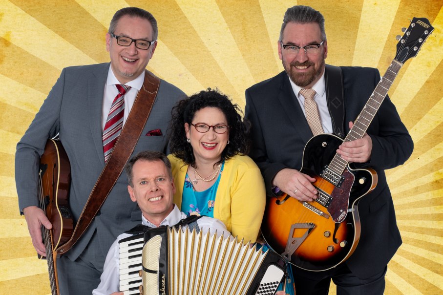 Four people pose cheerfully with instruments in front of a yellow ray background. Two men are holding guitars, one an accordion, a woman stands in the middle., © Friedrichsdbau Varieté Four people pose cheerfully with instruments in front of a yellow ray background. Two men are holding guitars, one an accordion, a woman stands in the middle., © Friedrichsdbau Varieté