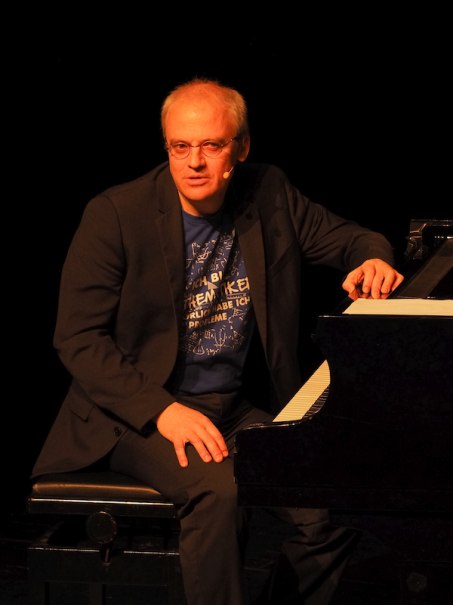 A man is sitting at a grand piano on a stage, wearing a microphone and a T-shirt with a mathematical print., &copy; Theaterhaus Stuttgart e.V.