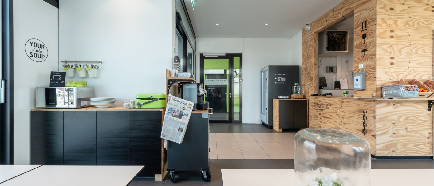 Moderne Hotel-Lounge mit Holzwänden, Snackbereich und Tisch mit Glasglocke. Zeitung und Kaffeemaschine sichtbar. Helle, einladende Atmosphäre., © a2 HOTELS