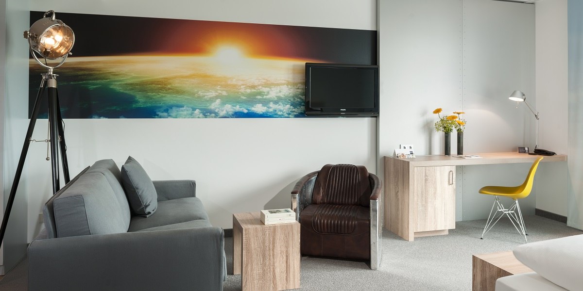 Modern hotel room with gray sofa, leather armchair, desk, yellow chair and large mural of a sunrise over the earth., &copy; Essential by Dorint