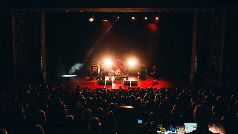 Eine Band spielt auf einer beleuchteten Bühne vor einem großen Publikum in einem dunklen Konzertsaal., © Württembergische Staatstheater Stuttgart Eine Band spielt auf einer beleuchteten Bühne vor einem großen Publikum in einem dunklen Konzertsaal., © Württembergische Staatstheater Stuttgart