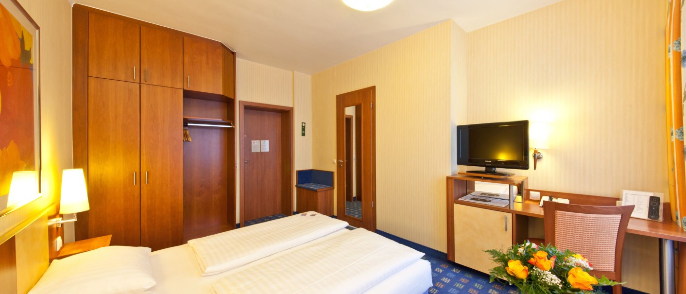 Ein gemütliches Hotelzimmer mit Doppelbett, Schreibtisch, Fernseher und einem Blumenstrauß auf einem Tisch., © Novum Boulevard Stuttgart City Ein gemütliches Hotelzimmer mit Doppelbett, Schreibtisch, Fernseher und einem Blumenstrauß auf einem Tisch., © Novum Boulevard Stuttgart City