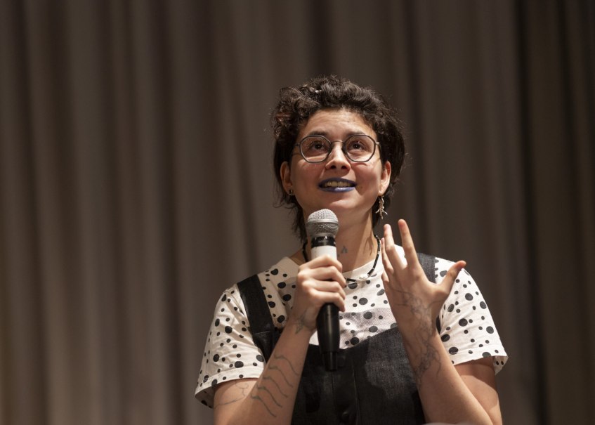 Eine Person mit Brille und blauem Lippenstift spricht in ein Mikrofon. Sie tr&auml;gt ein gepunktetes Oberteil und hat t&auml;towierte Arme., &copy; Astrid Ackermann