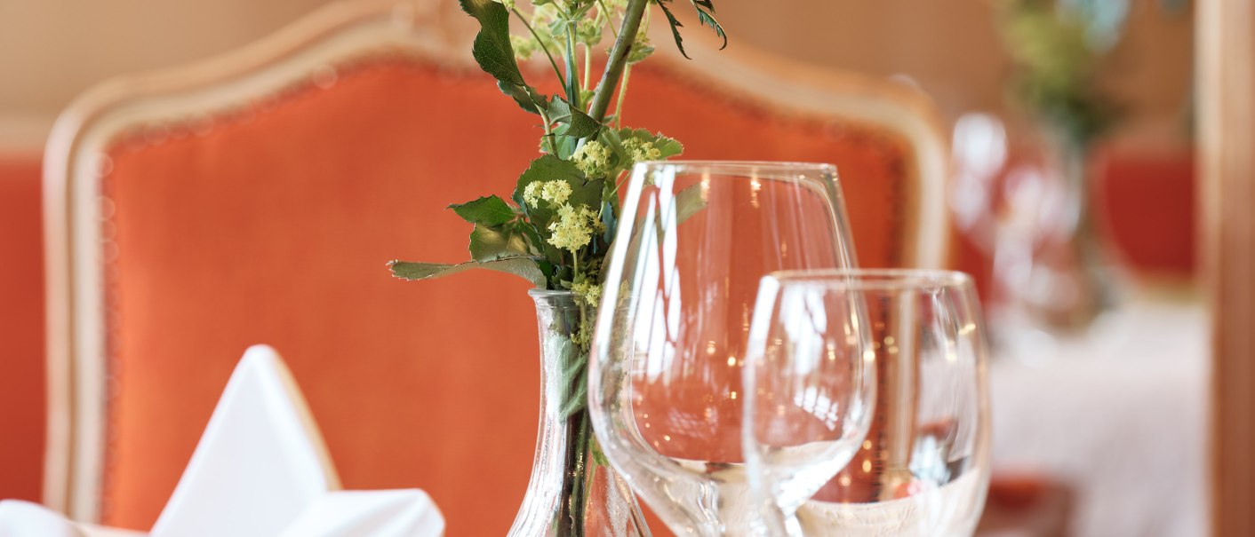 An elegant table with a yellow-and-red rose in a vase, wine glasses, and silverware on a white tablecloth, with an orange chair in the background., © Badhotel Restaurant Stauferland An elegant table with a yellow-and-red rose in a vase, wine glasses, and silverware on a white tablecloth, with an orange chair in the background., © Badhotel Restaurant Stauferland