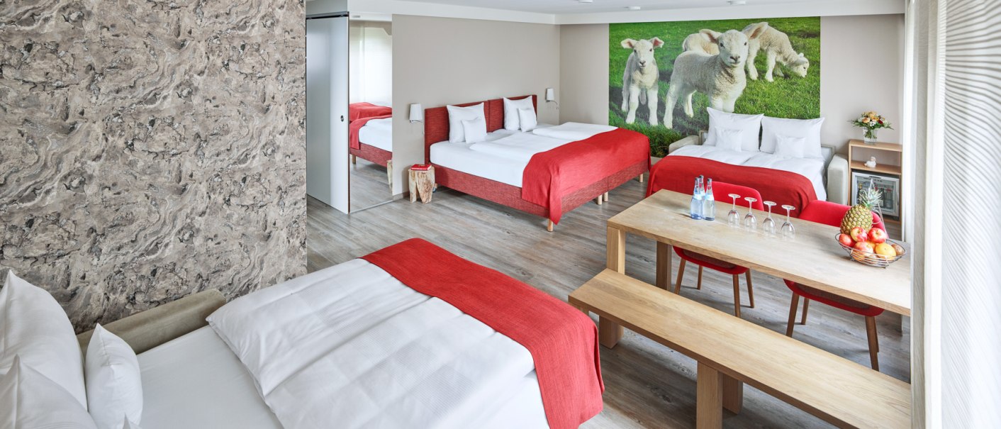 Geräumiges Hotelzimmer mit drei Betten, roter Bettwäsche, Holztisch und Wandbild von Lämmern. Modernes Design mit hellem Holzfußboden., © Hotel Restaurant Lamm Geräumiges Hotelzimmer mit drei Betten, roter Bettwäsche, Holztisch und Wandbild von Lämmern. Modernes Design mit hellem Holzfußboden., © Hotel Restaurant Lamm