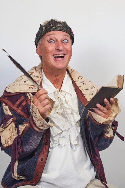 Person in bunter, historischer Kleidung mit Krone, hält eine Feder und ein Buch, lächelt breit in die Kamera., © Theaterhaus Stuttgart e.V.