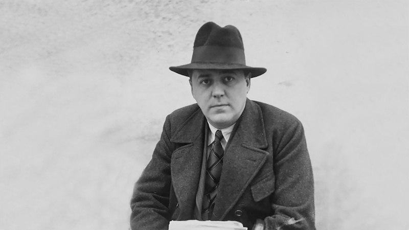 Ein Mann in einem Mantel und Hut sitzt mit ernstem Blick. Das Bild ist in Schwarz-Weiß gehalten., © tri-bühne e.V. Ein Mann in einem Mantel und Hut sitzt mit ernstem Blick. Das Bild ist in Schwarz-Weiß gehalten., © tri-bühne e.V.