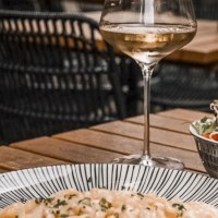 A glass of white wine, a plate of pasta and a salad on a wooden table in a restaurant with woven chairs., © SMG, itchyfeet-travel.de