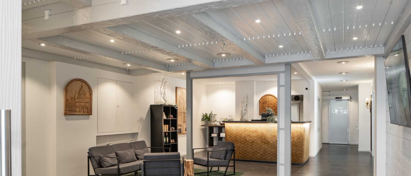 A modern reception area with a cozy seating area, wooden furniture, and a decorative ceiling. Bright lighting and plants create a welcoming atmosphere., &copy; Flairhotel Vier Jahreszeiten