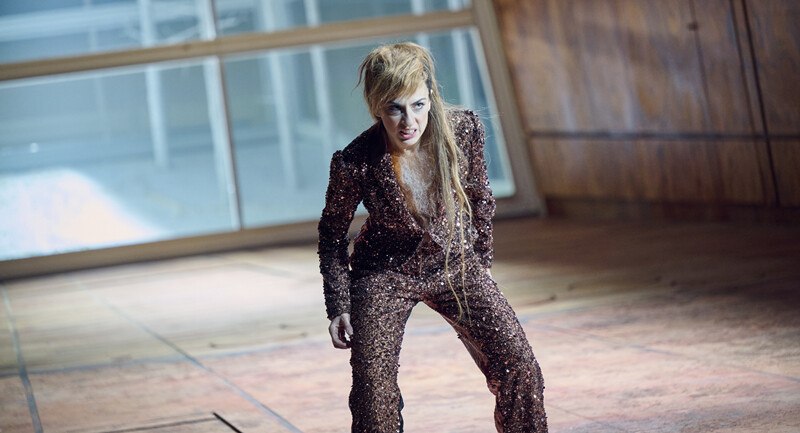 Person in glitzerndem Anzug auf Bühne, dramatische Pose. Hintergrund aus Holz und Glas., © Württembergische Staatstheater Stuttgart Person in glitzerndem Anzug auf Bühne, dramatische Pose. Hintergrund aus Holz und Glas., © Württembergische Staatstheater Stuttgart