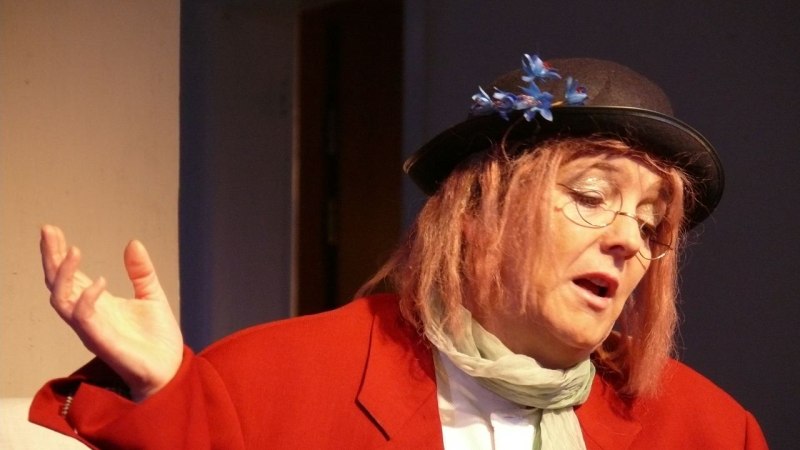 Eine Person in rotem Mantel und Hut mit blauen Blumen gestikuliert. Sie tr&auml;gt eine Brille und einen Schal., &copy; Stein im See_ Gesine Keller&copy;Foto: Katja Ritter