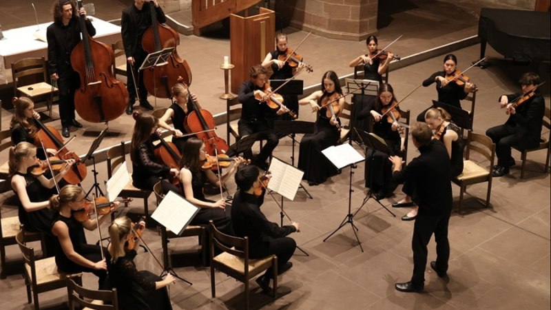 Ein Orchester mit Streichinstrumenten spielt in einem Konzertsaal. Die Musiker tragen schwarze Kleidung und werden von einer Person dirigiert., © JuKo (privat) Ein Orchester mit Streichinstrumenten spielt in einem Konzertsaal. Die Musiker tragen schwarze Kleidung und werden von einer Person dirigiert., © JuKo (privat)