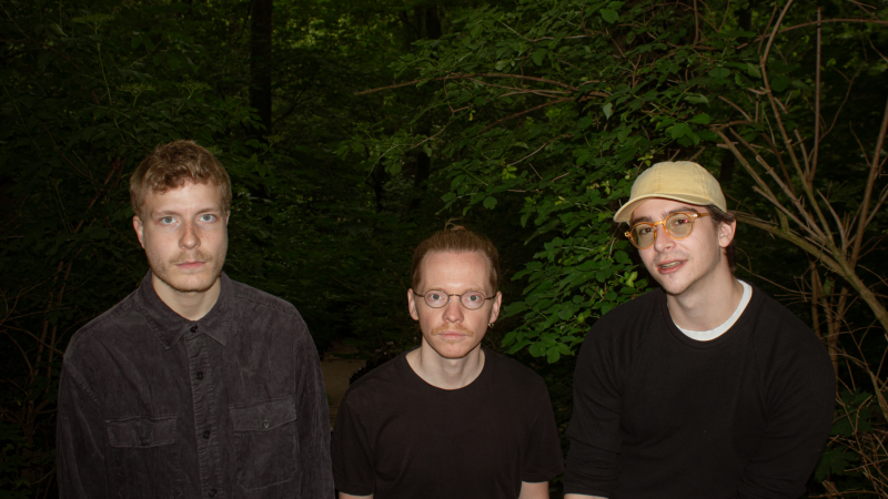 Drei Männer stehen im Wald, umgeben von dichtem Grün. Sie tragen dunkle Kleidung und schauen ernst in die Kamera., © Blanca Lopez Drei Männer stehen im Wald, umgeben von dichtem Grün. Sie tragen dunkle Kleidung und schauen ernst in die Kamera., © Blanca Lopez