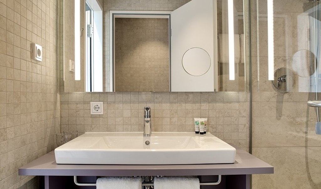 Modern bathroom with rectangular washbasin, illuminated mirror and shower area. Discreet tiles and towel rail under the washbasin., &copy; Hotel am Remspark
