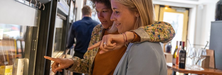Zwei Frauen stehen vor einem Weinspender und zeigen auf verschiedene Weinflaschen. Im Hintergrund sind weitere Flaschen und Gl&auml;ser zu sehen., &copy; Stuttgart Marketing GmbH, Fotografin Martina Denker