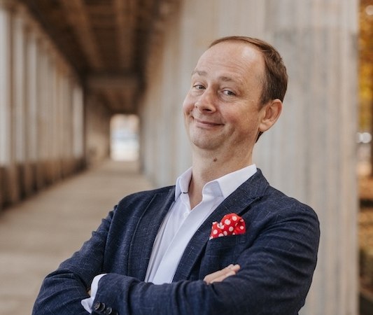 A man in a blue suit with a red handkerchief stands smiling in a portico., &copy; Renitenztheater Stuttgart e.V.