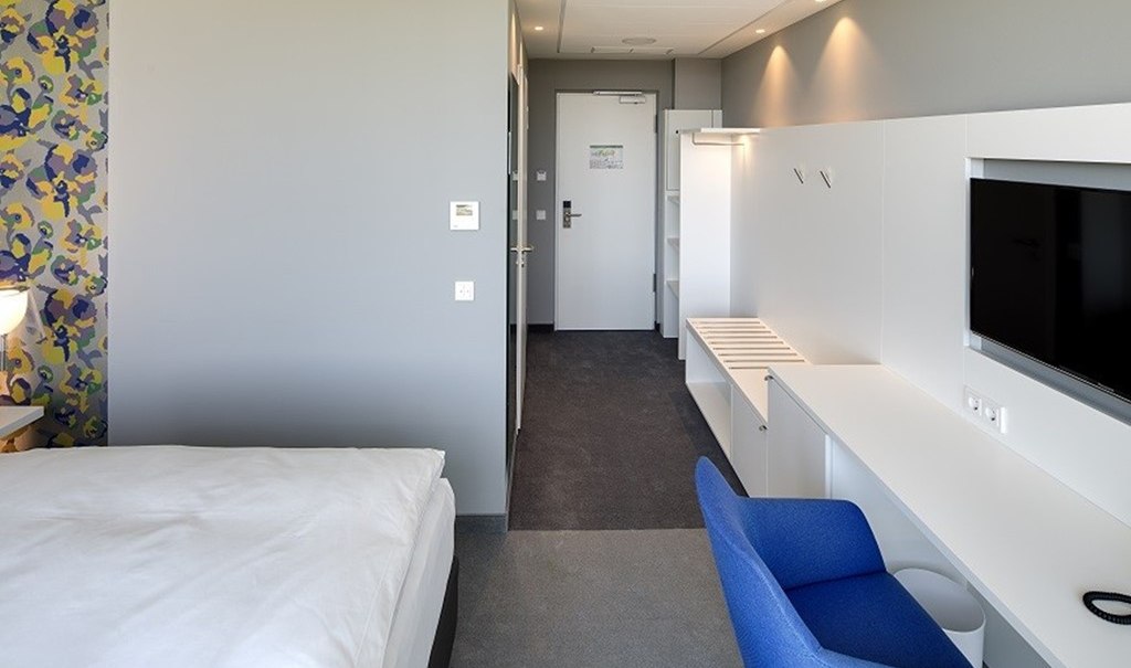 Modern single room with bed, desk, blue chair and TV. Bright walls and colorful wallpaper on one side., © Hotel am Remspark Modern single room with bed, desk, blue chair and TV. Bright walls and colorful wallpaper on one side., © Hotel am Remspark
