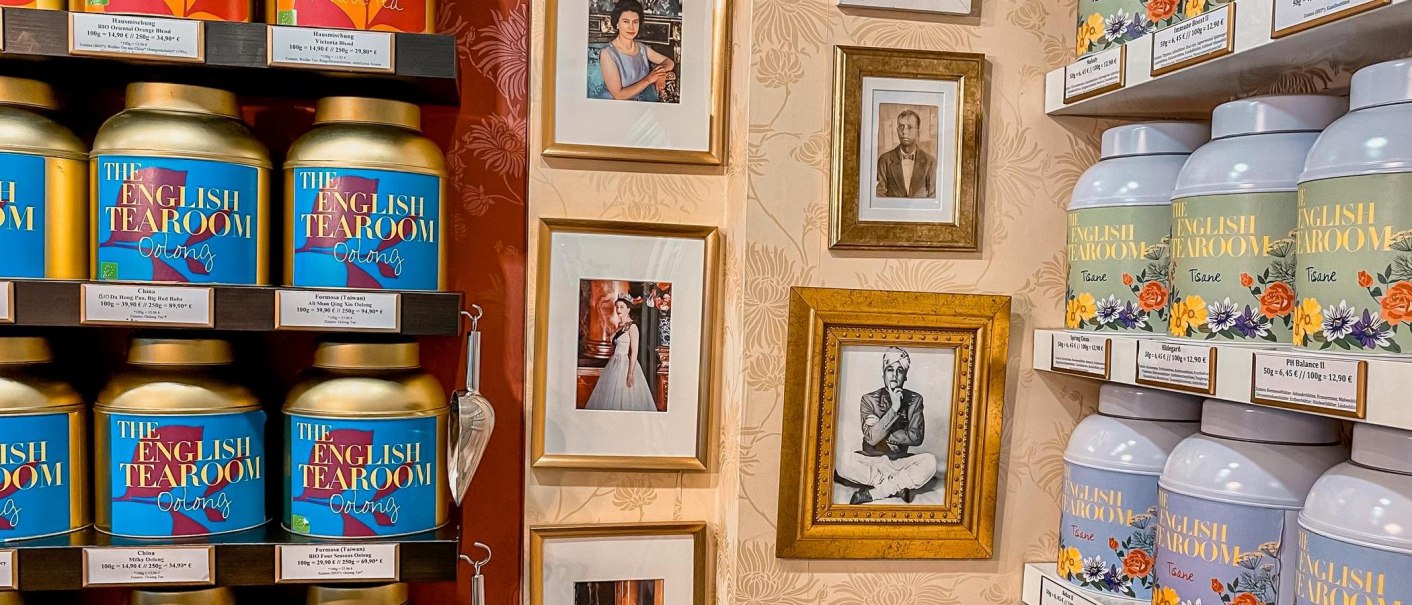 Regale mit Teedosen von 'The English Tearoom' und gerahmte Fotos an einer dekorativen Wand., &copy; Tea Room, Stuttgart