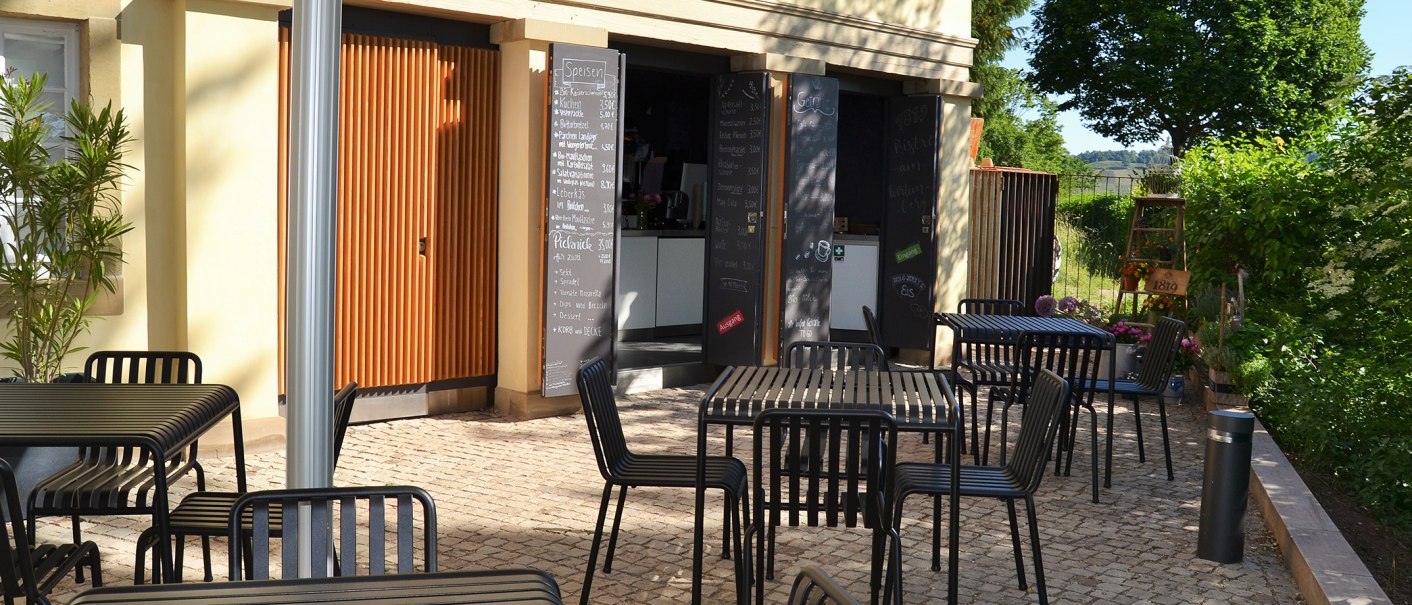 Outdoor area of the 1819 Bistro am Wirtemberg with black tables and chairs on paved ground, surrounded by green vegetation., © 1819 Bistro am Wirtemberg