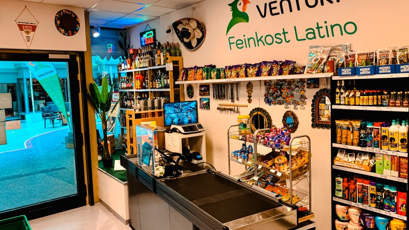 Ein Feinkostladen mit lateinamerikanischen Produkten, bunten Dekorationen und einer Kasse. Regale sind mit Snacks und Getränken gefüllt., © Feinkost Latino Ventura