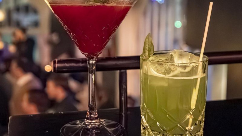 Zwei Cocktails auf einer Bar: ein roter im Martiniglas und ein grüner im Tumblerglas mit Strohhalm, im Hintergrund unscharfe Bar-Szene., © SMG, Sarah Schmid