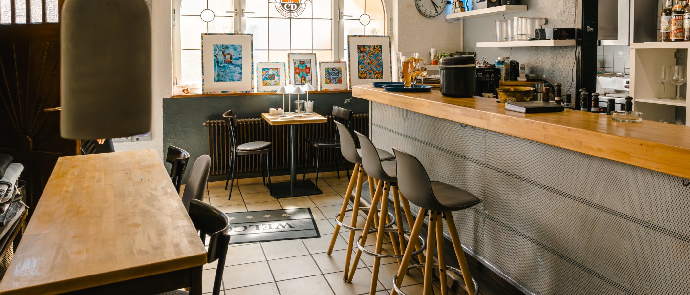 Gemütlicher Café-Innenraum mit Theke, Barhockern und dekorativen Bildern. Große Fenster lassen viel Licht herein. Eine Uhr und ein "Open"-Schild sind sichtbar., © SMG Stuttgart Marketing GmbH - Sarah Schmid
