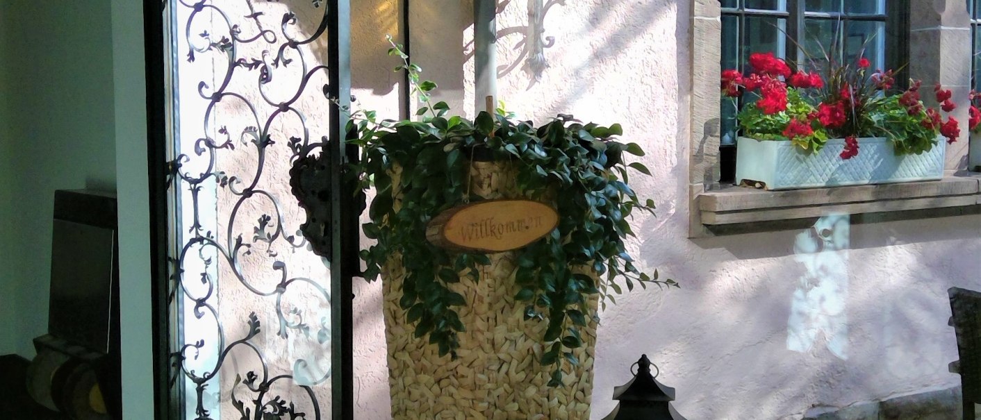 An inviting entrance with an open wrought-iron gate, a large plant basket with a 'Welcome' sign, lanterns and a flower box by the window., &copy; relexa Waldhotel Schatten
