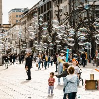 Ein Straßenkünstler erzeugt Seifenblasen auf einer belebten Straße. Kinder spielen begeistert mit den Blasen, während Passanten zuschauen., © Stuttgart-Marketing GmbH, Sarah Schmid Ein Straßenkünstler erzeugt Seifenblasen auf einer belebten Straße. Kinder spielen begeistert mit den Blasen, während Passanten zuschauen., © Stuttgart-Marketing GmbH, Sarah Schmid
