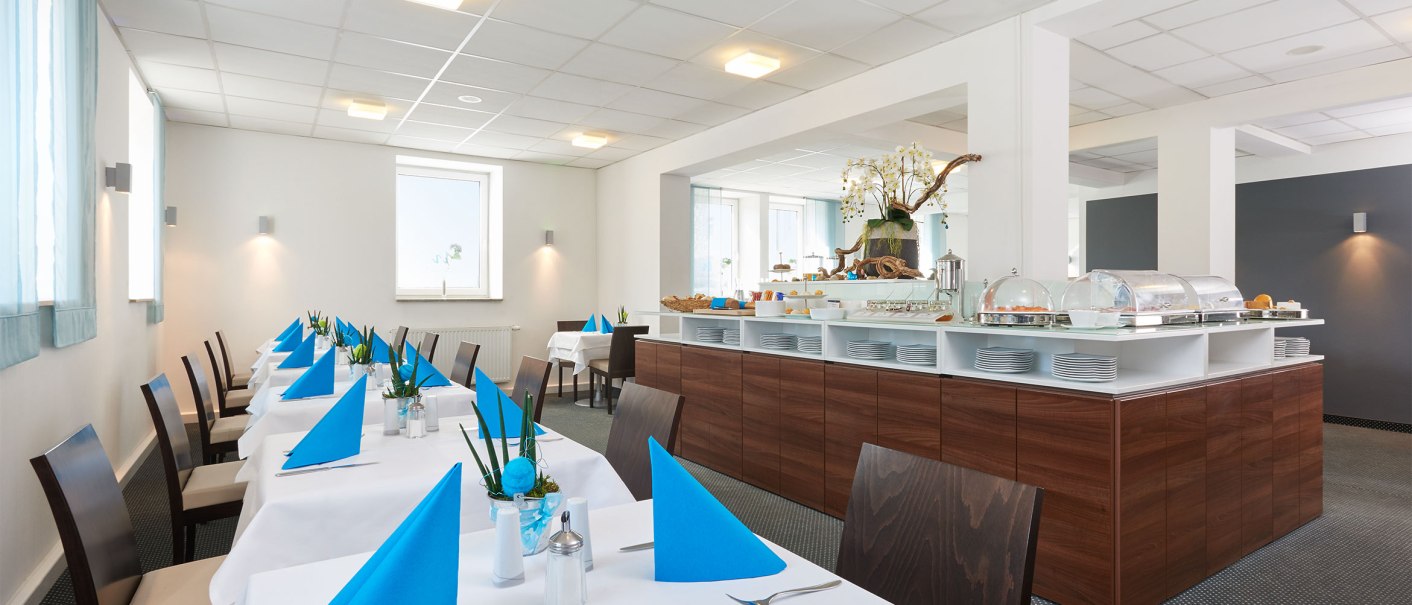 Ein heller Frühstücksraum mit weiß gedeckten Tischen, blauen Servietten und einem Buffet im Hintergrund., © TOMAS Ein heller Frühstücksraum mit weiß gedeckten Tischen, blauen Servietten und einem Buffet im Hintergrund., © TOMAS