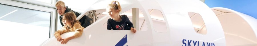 Drei Kinder spielen in einem Cockpit-Nachbau mit der Aufschrift 'SkyLand Cockpit' und blicken aus den Fenstern. Helle, moderne Umgebung., © Flughafen Stuttgart