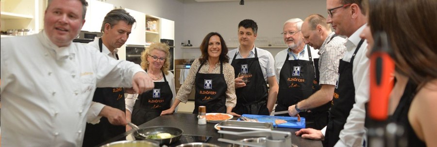 Menschen in einer K&uuml;che tragen 'Ilzh&ouml;fer's' Sch&uuml;rzen und kochen gemeinsam. Ein Koch zeigt etwas in einer Pfanne., &copy; Ilzh&ouml;fer, Stuttgart