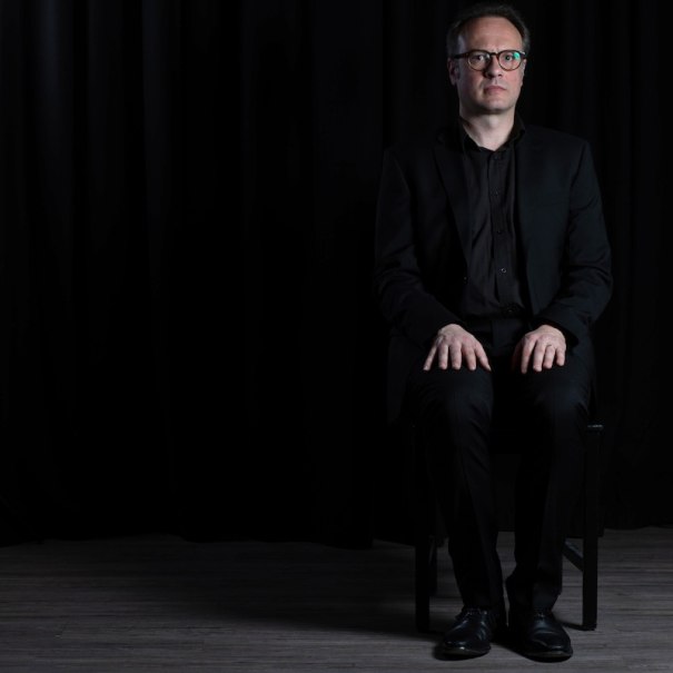 Person in a black suit sits on a chair in front of a black background, looks seriously into the camera., © Renitenztheater Stuttgart e.V. Person in a black suit sits on a chair in front of a black background, looks seriously into the camera., © Renitenztheater Stuttgart e.V.