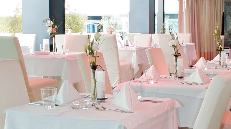 Elegantes Restaurant mit weißen Tischdecken, stilvoll gedeckten Tischen und Blumenarrangements. Helle, moderne Einrichtung mit großen Fenstern., © AMICI