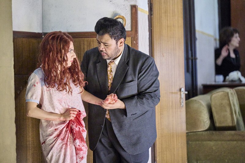 Ein Mann im Anzug hält eine Frau in einem blutbefleckten Kleid. Im Hintergrund ist eine weitere Person zu sehen., © Württembergische Staatstheater Stuttgart