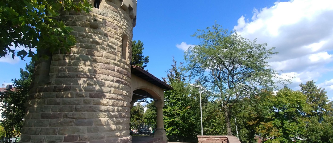 Runder Steinturm mit Zinnen, umgeben von Bäumen und blauem Himmel. Der Turm steht auf einer gepflasterten Fläche., © SMG Runder Steinturm mit Zinnen, umgeben von Bäumen und blauem Himmel. Der Turm steht auf einer gepflasterten Fläche., © SMG