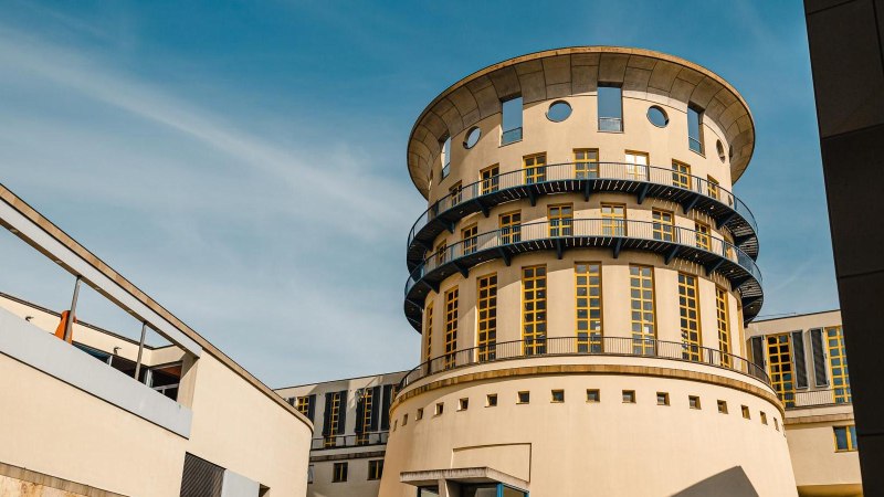 Rundes Gebäude mit modernen Elementen und blauen Balkonen, umgeben von weiteren Gebäudeteilen, unter blauem Himmel., © Stuttgart-Marketing GmbH, Sarah Schmid