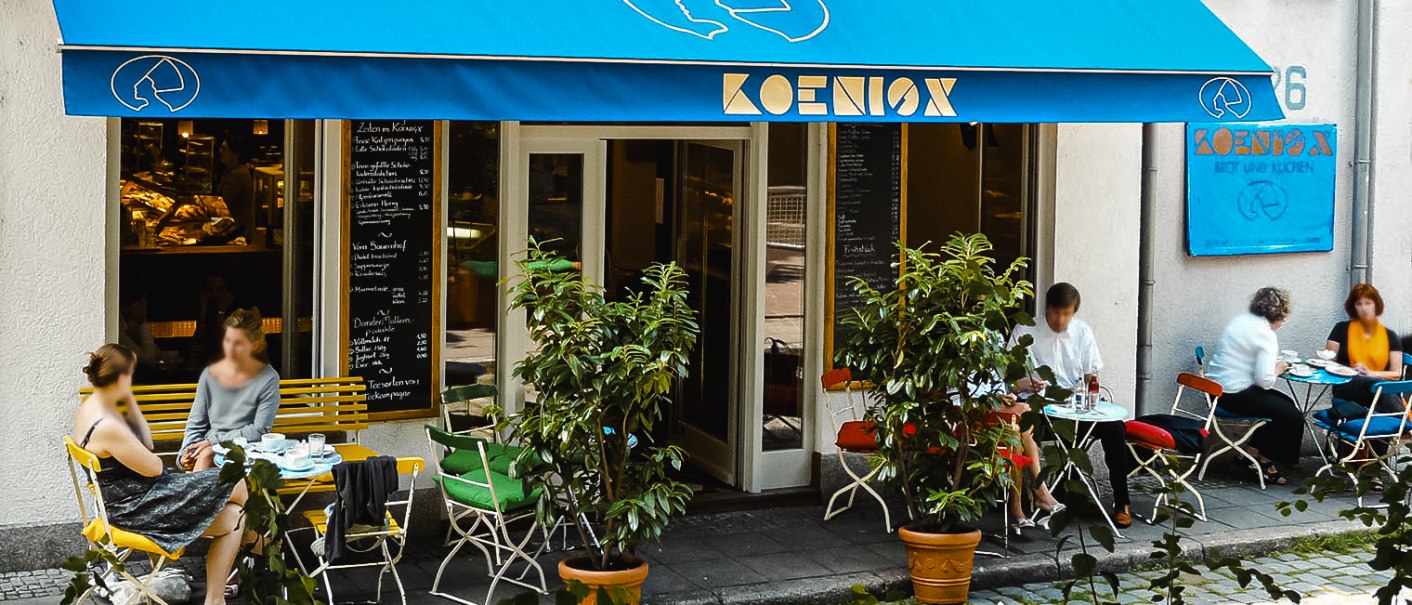 Ein Café mit blauer Markise und dem Namen 'Koenig'. Gäste sitzen draußen an Tischen und genießen ihre Getränke. Pflanzen dekorieren den Eingangsbereich., © KoenigX