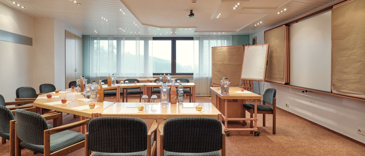 A bright seminar room with U-shaped tables, chairs, flip charts, and drinks on the tables. Large windows let in natural light., © Badhotel Restaurant Stauferland A bright seminar room with U-shaped tables, chairs, flip charts, and drinks on the tables. Large windows let in natural light., © Badhotel Restaurant Stauferland