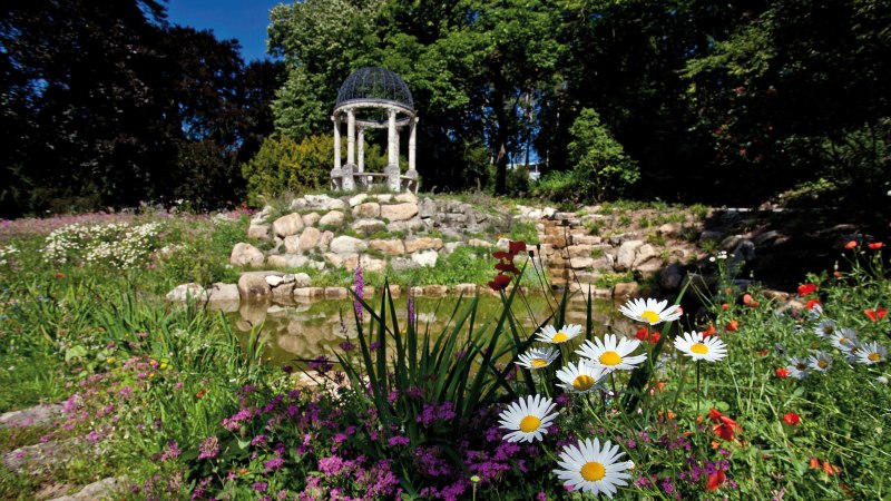 Ein blühender Garten mit bunten Blumen, einem kleinen Teich und einem Pavillon im Hintergrund, umgeben von Bäumen., © Staatsministerium Baden-Württemberg Ein blühender Garten mit bunten Blumen, einem kleinen Teich und einem Pavillon im Hintergrund, umgeben von Bäumen., © Staatsministerium Baden-Württemberg