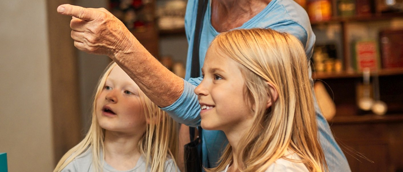 An older woman shows two young girls something in a museum. The girls look with interest in the direction the woman is pointing., © FTGRF.de An older woman shows two young girls something in a museum. The girls look with interest in the direction the woman is pointing., © FTGRF.de
