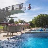 Ein Freibad mit Sprungturm, von dem ein Mensch ins Wasser springt. Viele Menschen schwimmen im großen Becken. Im Hintergrund sind Bäume und Sonnenschirme., © Stuttgarter Bäder Ein Freibad mit Sprungturm, von dem ein Mensch ins Wasser springt. Viele Menschen schwimmen im großen Becken. Im Hintergrund sind Bäume und Sonnenschirme., © Stuttgarter Bäder