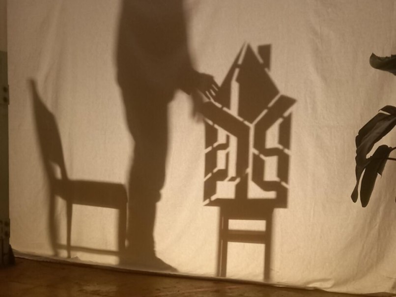 Ein Schattenspiel zeigt die Silhouette eines Menschen, der ein Haus auf einem Stuhl berührt. Die Szene ist auf eine Leinwand projiziert., © Esther Falk Ein Schattenspiel zeigt die Silhouette eines Menschen, der ein Haus auf einem Stuhl berührt. Die Szene ist auf eine Leinwand projiziert., © Esther Falk