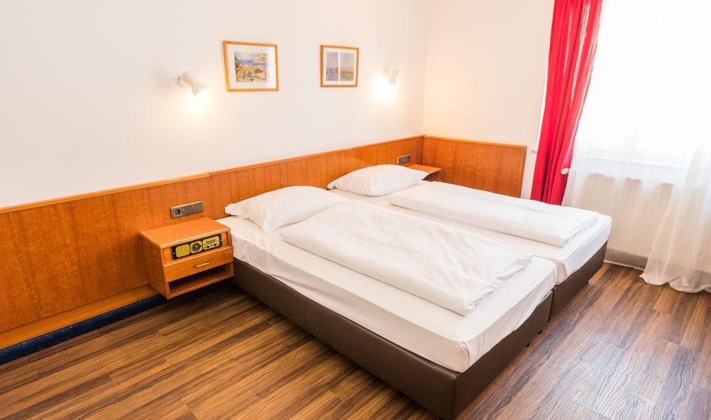 Ein Doppelzimmer mit zwei Einzelbetten, Holzboden, roten Vorhängen und zwei Bildern an der Wand., © ARNOTEL GmbH