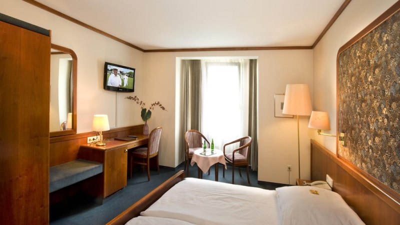 Gemütliches Hotelzimmer mit Doppelbett, Fernseher, Schreibtisch und Sitzbereich vor einem Fenster. Holzmöbel und warme Beleuchtung dominieren den Raum., © Hotel am Schelztor Gemütliches Hotelzimmer mit Doppelbett, Fernseher, Schreibtisch und Sitzbereich vor einem Fenster. Holzmöbel und warme Beleuchtung dominieren den Raum., © Hotel am Schelztor