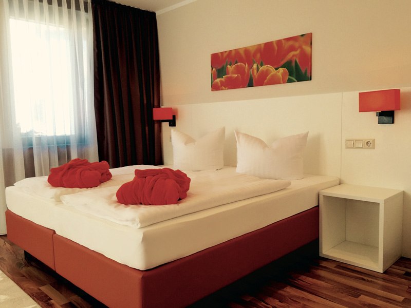 Modernes Hotelzimmer mit Doppelbett, roten Akzenten, Bild von Blumen an der Wand und zwei roten Badem&auml;nteln auf dem Bett., &copy; TOMAS