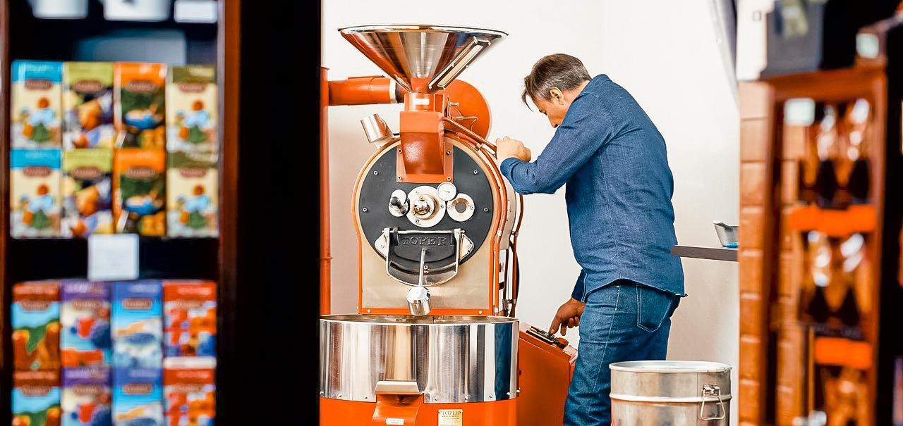 Ein Mann in Jeans bedient eine große, orangefarbene Kaffeeröstmaschine. Im Hintergrund sind bunte Kaffeepackungen zu sehen., © Kaffee Fröhlich, Stuttgart