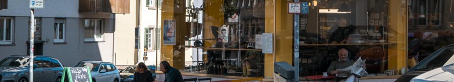 Ein Caf&eacute; namens Kantinchen mit gelbem und gr&uuml;nem Schild. Drau&szlig;en sitzen Menschen an bunten Tischen. Verkehrsschilder und Autos sind zu sehen., &copy; SMG, Sarah Schmid